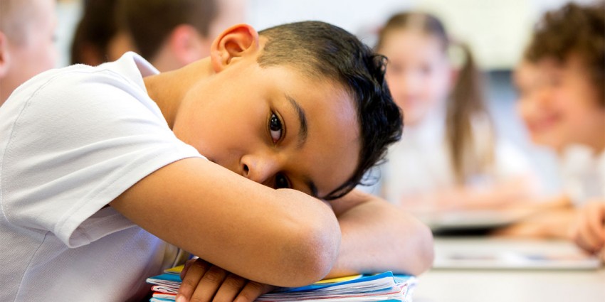 A young boy in school with his head on his desk, looking sad.