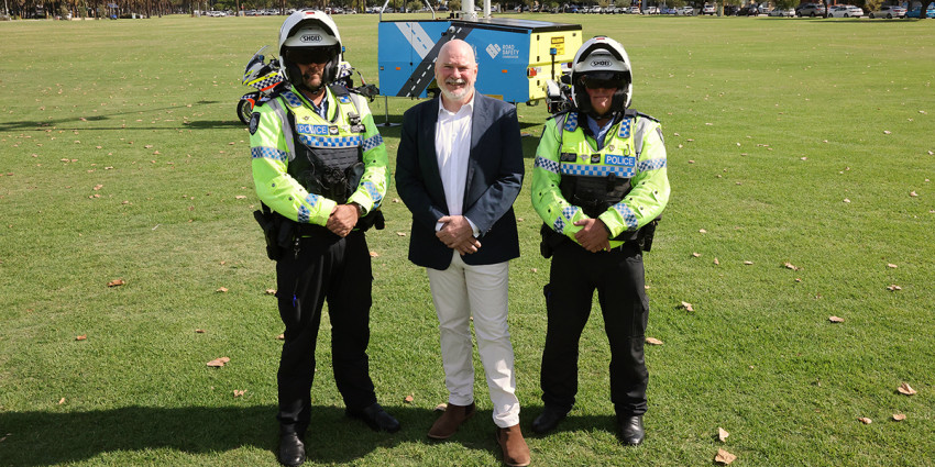 Road Safety Commissioner Adrian Warner flanked by two police officers standing in a field in front of a safety camera.